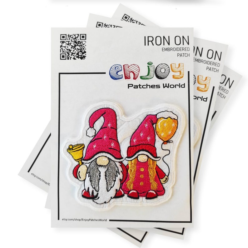 K&ouml;nnte beinhalten: Ein aufb&uuml;gelbarer gestickter Patch mit zwei Gnomen. Ein Gnom h&auml;lt eine Glocke und der andere einen Ballon. Die Gnome tragen rote M&uuml;tzen und rote Hemden. Der Patch ist wei&szlig; mit einem roten und wei&szlig;en Schneeflockenmuster. Der Text "IRON ON" und "EMBROIDERED PATCH" ist auf der Verpackung aufgedruckt. Der Text "enjoy Patches World" ist unter den Gnomen aufgedruckt.