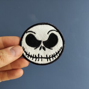 Könnte beinhalten: Ein gestickter Aufnäher mit dem Gesicht von Jack Skellington aus dem Film Nightmare Before Christmas. Der Aufnäher ist weiß mit schwarzen Nähten.