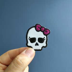 Puede incluir: Cráneo blanco con ojos negros y un lazo rosa. El cráneo está bordado en una tela blanca.