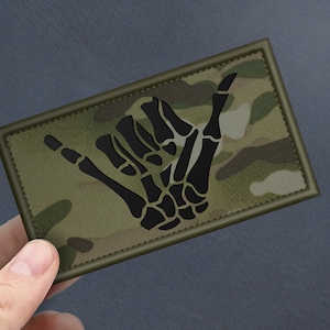 Pode incluir: Um remendo de tecido camuflado verde, marrom e bege com uma mão de esqueleto preta fazendo o sinal shaka, um gesto popular havaiano.
