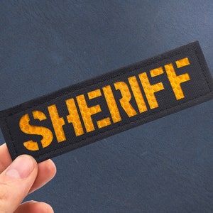 May include: A black fabric patch with the word "SHERIFF" in yellow reflective lettering.