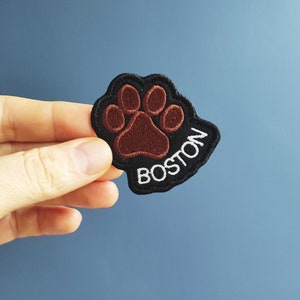 Könnte beinhalten: Gestickter Aufnäher mit braunem Pfotenabdruck auf schwarzem Grund. Das Wort "BOSTON" ist weiß gestickt. Der Aufnäher wird vor einem blauen Hintergrund gehalten.