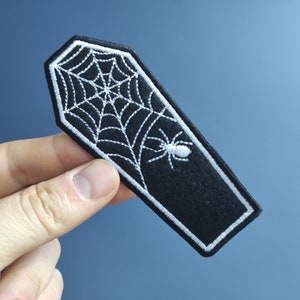 Könnte beinhalten: Schwarzer Filz-Patch in Sargform mit weiß gesticktem Spinnennetz und Spinne.