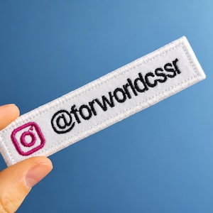 May include: A rectangular white embroidered patch featuring a magenta Instagram icon on the left side and the black text @forworldcssr embroidered on the right side, with a white stitched border surrounding the entire rectangular fabric piece.