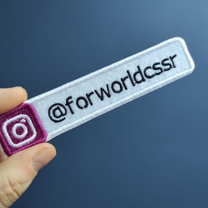 Könnte beinhalten: Weißer Stoff-Patch mit schwarzem gesticktem Text "@forworldcssr" und einem rosafarbenen Instagram-Logo.