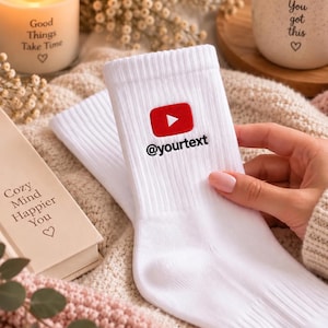 Könnte beinhalten: Ein Paar weiße, gerippte Socken mit einem roten YouTube-Wiedergabetaste-Symbol und dem schwarzen Text @yourtext, der auf den oberen Bund gestickt ist. Die Socken bestehen aus einem weichen Strickstoff und haben ein schlichtes, klares Design.