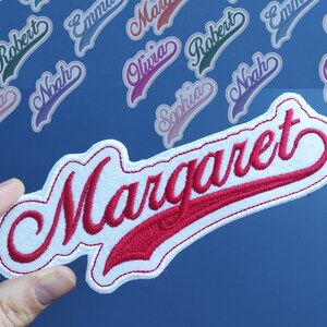 Pode incluir: Remendo de feltro branco com letras bordadas em vermelho que dizem "Margaret" em uma fonte cursiva. O remendo tem a forma de uma camisa de beisebol.