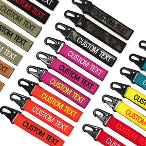 Può includere: Una collezione di portachiavi con testo personalizzato in vari colori, tra cui rosso, rosa, giallo, verde e nero. Ogni portachiavi presenta un moschettone nero e le parole "CUSTOM TEXT" in lettere bianche o gialle.