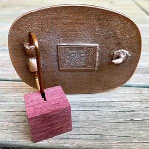 May include: A bronze-coloured metal belt buckle with a rectangular stamp that reads "PEE GOOD". A small, square, purple wooden block is attached to the buckle. The buckle is oval-shaped.