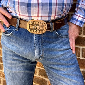 May include: A brown leather belt with a brass buckle featuring the text "Ladies My Eyes Are Up Here". The belt is worn with blue denim jeans and a plaid shirt. The buckle is the central element of the image.