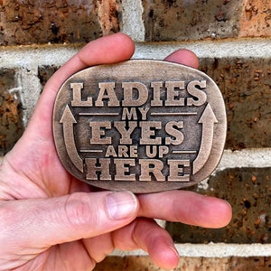 May include: Bronze-coloured metal belt buckle with the text "LADIES MY EYES ARE UP HERE" in bold, capital letters. The buckle has an oval shape with an arrow design. The background is a brick wall.