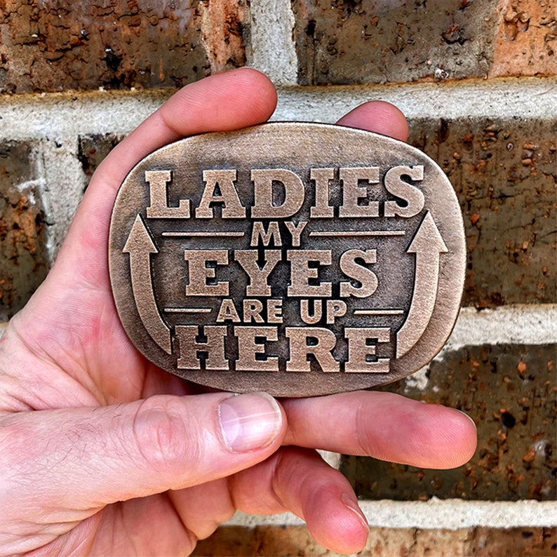 May include: Bronze-coloured metal belt buckle with the text "LADIES MY EYES ARE UP HERE" in bold, capital letters. The buckle has an oval shape with an arrow design. The background is a brick wall.