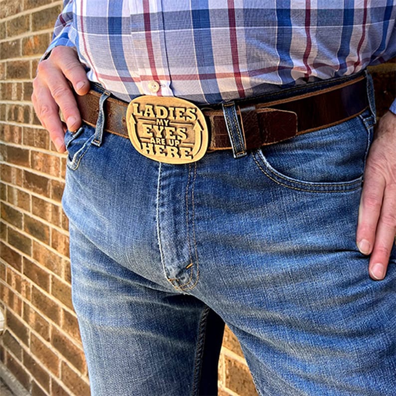 May include: A brown leather belt with a gold-coloured buckle. The buckle features the text "LADIES MY EYES ARE UP HERE". The belt is worn with blue jeans and a plaid shirt. The buckle is a decorative accessory.