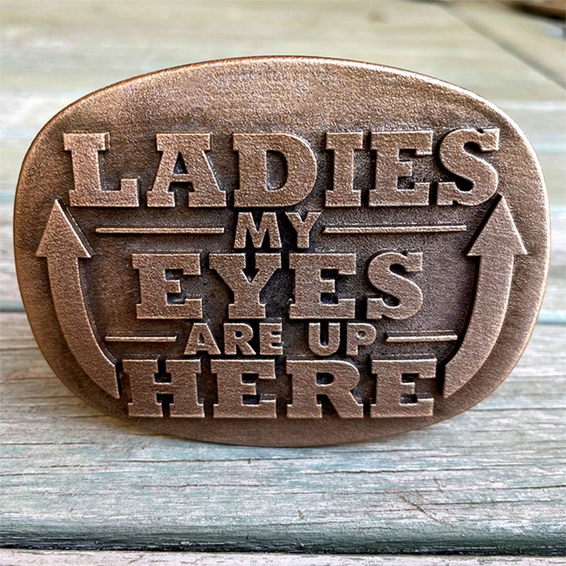 May include: Bronze-coloured oval belt buckle with the text "LADIES MY EYES ARE UP HERE" in raised lettering. The buckle has an arrow design on each side, pointing upwards. The buckle is on a light blue wooden surface.