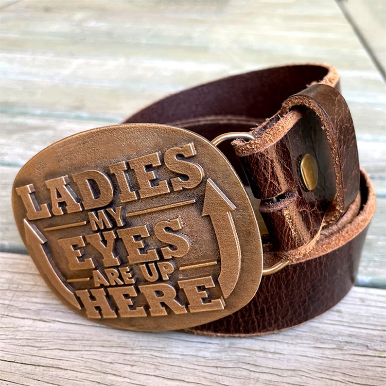 May include: A brown leather belt featuring a bronze buckle. The buckle is inscribed with the words "LADIES MY EYES ARE UP HERE" and an upward-pointing arrow. The belt is made of distressed leather.
