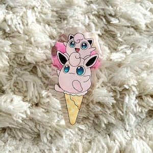 Może przedstawiać: Różowo-biała postać Pokemon, Jigglypuff, przedstawiona jest w wafelku lodowym. Postać trzyma na głowie mniejszego Jigglypuffa. Wafel lodowy jest żółty i biały.