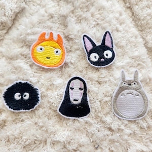 May include: Five embroidered iron-on patches featuring characters from popular Japanese animated films. The patches are on a white fluffy background. The patches include a black cat, a white and grey creature with large ears, a black and white creature with a face, a yellow and orange creature with a face, and a black and white creature with a face.