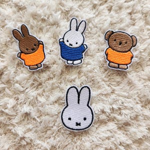 May include: Four embroidered patches of cartoon characters. Two are brown bunnies in orange tops, one is a white bunny in a blue top, one is a brown bear in an orange top, and one is a white bunny with a black outline.