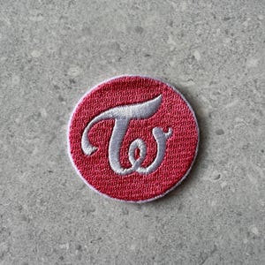 May include: A circular embroidered patch featuring a vibrant red background with a white stitched border. Centered on the patch is a stylized white letter T and a lowercase w intertwined in a cursive script font, creating a logo design.