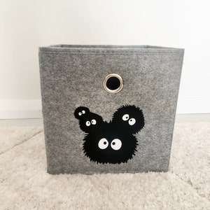 May include: A gray felt storage bin with a black and white graphic of a cartoon character with large eyes. The bin has a metal ring handle on the top.