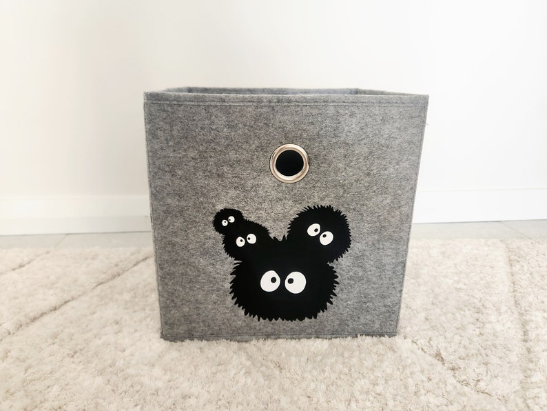 May include: A gray felt storage bin with a black and white graphic of a cartoon character with large eyes. The bin has a metal ring handle on the top.