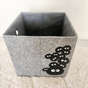 May include: A gray felt storage bin with a black and white graphic of a cartoon character with multiple eyes. The bin has a round hole on the side.