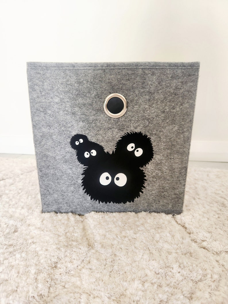 May include: A gray felt storage bin with a black and white graphic of a cartoon character with large eyes. The bin has a black metal handle.