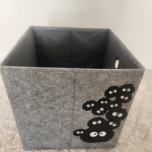 May include: A gray felt storage box with a black and white graphic of a group of Totoro characters. The box has a round hole on the side.