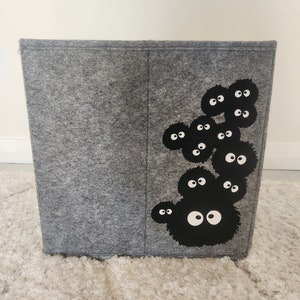 May include: A gray felt storage bin with a black and white graphic of a group of cartoon characters with large eyes. The characters are arranged in a cluster on the right side of the bin.