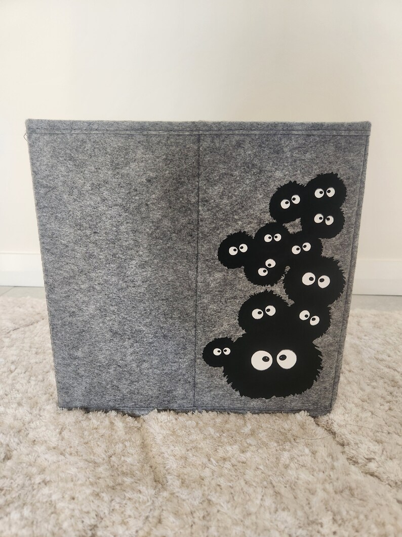 May include: A gray felt storage bin with a black and white graphic of a group of cartoon characters with large eyes. The characters are arranged in a cluster on the right side of the bin.