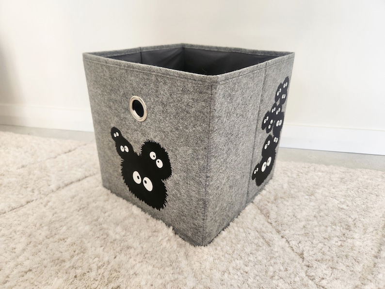 May include: A gray felt storage bin with a black and white graphic of a cartoon character with large eyes. The bin has a black handle on the top.