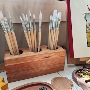 Puede incluir: Un soporte de pinceles de madera con tres compartimentos, cada uno lleno de una variedad de pinceles blancos. El soporte está hecho de madera marrón claro con un acabado natural.