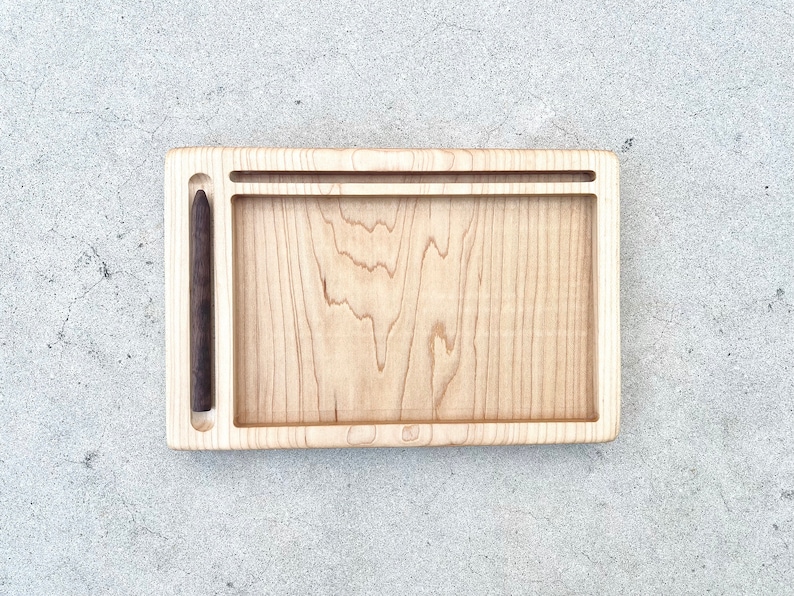 May include: A light wood tray with a dark wood groove for a pen or pencil. The tray is rectangular and has a smooth surface.