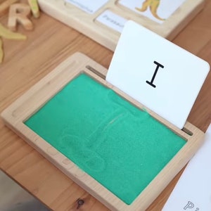 May include: A wooden tray with a green sand tray and a white card with the letter "I" on it. The card is propped up against the tray.