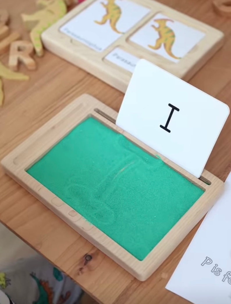 May include: A wooden tray with a green sand tray and a white card with the letter "I" on it. The card is propped up against the tray.