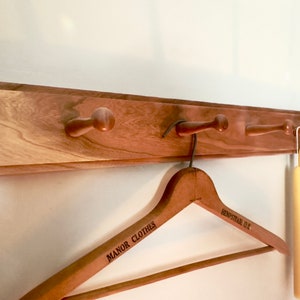 May include: Wooden wall-mounted coat rack with four pegs. A wooden clothes hanger with the text "MANOR CLOTHES HEMPSTEAD D.C." hangs from one peg. Two yellow candles hang from another peg.