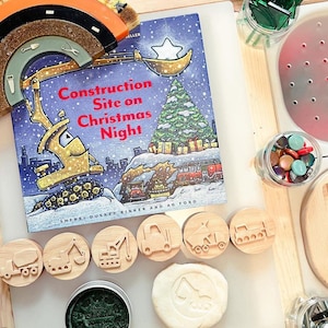 May include: A children's book titled "Construction Site on Christmas Night" by Sherri Duskey Rinker and AG Ford, featuring a yellow construction vehicle, a Christmas tree, and a star. The book is open on a table with wooden stamps of construction vehicles and a white play dough with a construction vehicle imprint.