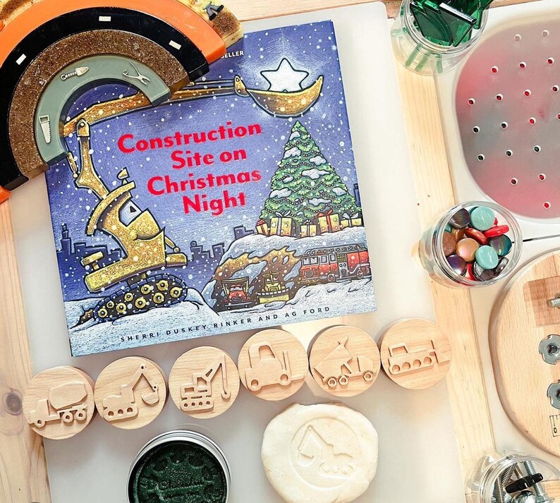 May include: A children's book titled "Construction Site on Christmas Night" by Sherri Duskey Rinker and AG Ford, featuring a yellow construction vehicle, a Christmas tree, and a star. The book is open on a table with wooden stamps of construction vehicles and a white play dough with a construction vehicle imprint.
