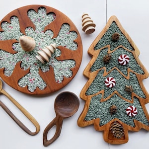 May include: Two wooden trays filled with rice and decorated with Christmas-themed cutouts. One tray is shaped like a snowflake and the other is shaped like a Christmas tree. The snowflake tray has red and white candy canes and the Christmas tree tray has red and white candy canes, pine cones, and small wooden ornaments.