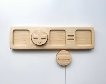 May include: Wooden tray with four compartments, two of which are empty. The middle compartment has a plus sign carved into it. The rightmost compartment has an equals sign carved into it. A separate wooden circle with a minus sign carved into it is below the tray.
