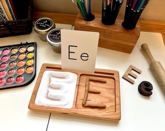May include: A wooden tray with two compartments for learning the letter 'E'. One compartment has a white powder for tracing the letter, and the other compartment has a brown dough for forming the letter. A white card with the letter 'Ee' is displayed on the tray.
