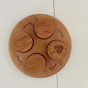 Peut inclure: Un plateau circulaire en bois avec quatre cercles encastr&eacute;s, chacun pr&eacute;sentant un motif diff&eacute;rent. Les motifs comprennent une grenouille, un poisson, un n&eacute;nuphar et une libellule. Le plateau est en bois brun clair et a une finition naturelle.