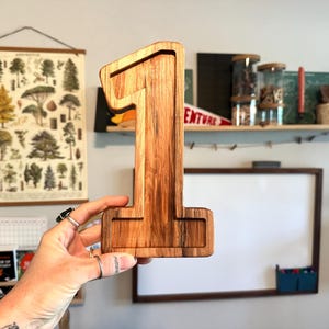 Könnte beinhalten: Eine hölzerne Nummer eins, in hellbraun lackiert, mit einer leicht dunkleren braunen Holzmaserung. Die Nummer wird in der Hand einer Person gehalten.