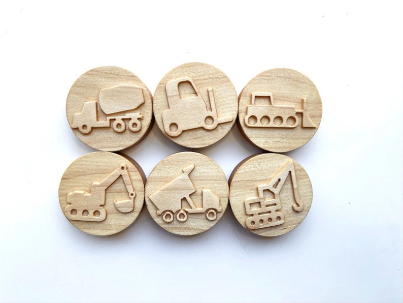 May include: Six wooden stamps featuring different construction vehicles. The stamps are round and have a light brown color. The vehicles include a cement mixer, a forklift, a bulldozer, an excavator, a dump truck, and a crane.