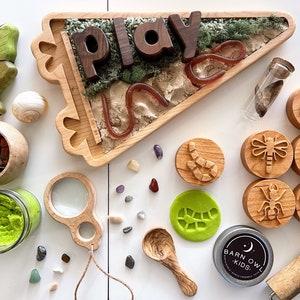 May include: A wooden tray with the word "play" spelled out in wooden letters, surrounded by green moss and a red worm. The tray is on a white surface with a variety of other items, including a magnifying glass, a jar of green play dough, a wooden spoon, and a set of wooden stamps with insect and worm designs.