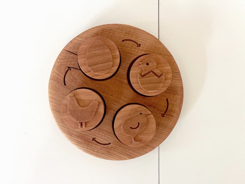 Peut inclure: Puzzle circulaire en bois compos&eacute; de quatre pi&egrave;ces. Les pi&egrave;ces repr&eacute;sentent un poussin &eacute;closant d'un &oelig;uf, un poussin et un &oelig;uf. Le puzzle est en bois brun clair.