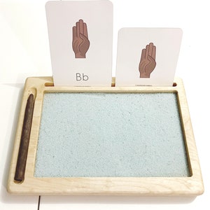 May include: A wooden tray with a sand-filled area for writing practice. Two cards with images of hands and the letter "Bb" are displayed on the tray.