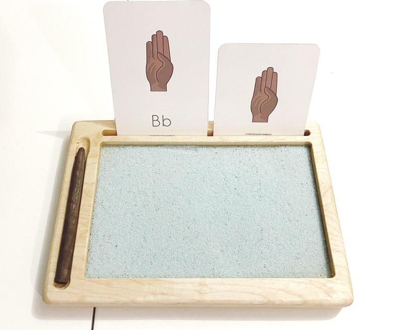 May include: A wooden tray with a sand-filled area for writing practice. Two cards with images of hands and the letter "Bb" are displayed on the tray.