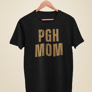 Könnte beinhalten: Schwarzes T-Shirt mit dem Text "PGH MOM" in goldenen Buchstaben.