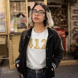 May include: A woman wearing a white t-shirt with the number "412" printed in gold on the front. She is also wearing a black jacket and blue jeans.